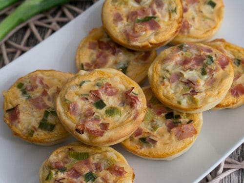 Mini-Quiches mit Schinken und Frühlingszwiebeln auf einem weißen Teller.