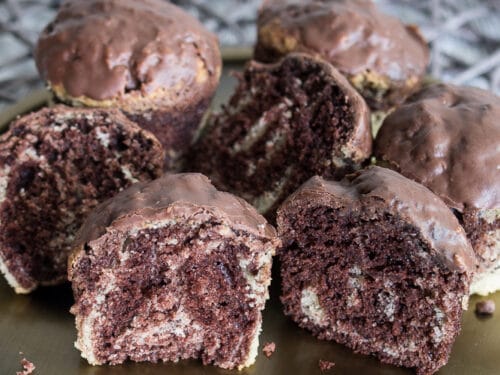 Sechs Marmorkuchen Muffins mit Schokoladenglasur auf einem Teller, einige aufgeschnitten, um ihr marmoriertes Innere zu zeigen.