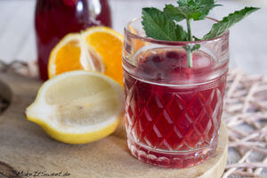 Ein Glas Sommer Eistee mit Minze und Beeren, begleitet von Zitronen- und Orangenscheiben auf einem Holzbrett.