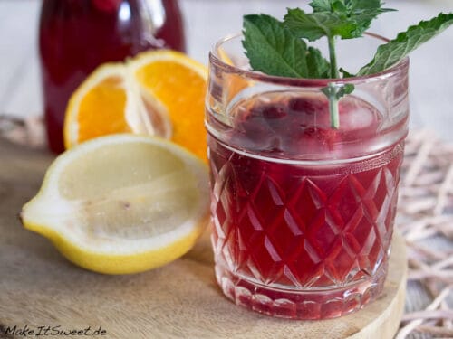 Ein Glas Sommer Eistee mit Minze und Beeren, begleitet von Zitronen- und Orangenscheiben auf einem Holzbrett.