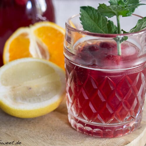 Sommer Eistee 3 Ein Glas Sommer Eistee mit Minze und Beeren, begleitet von Zitronen- und Orangenscheiben auf einem Holzbrett.