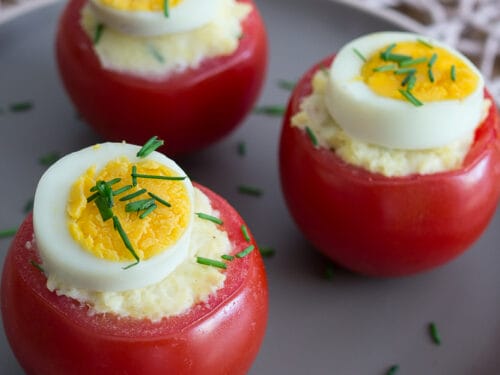 Drei mit Kartoffeln gefüllte Tomaten, belegt mit Eierscheiben und Schnittlauch, angerichtet auf einem grauen Teller.