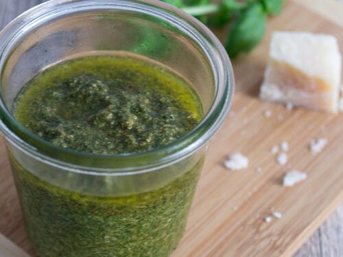 Ein Glas Basilikum-Pesto selbst gemacht mit frischen Basilikumblättern und einem Stück Parmesan auf einem Holzbrett.