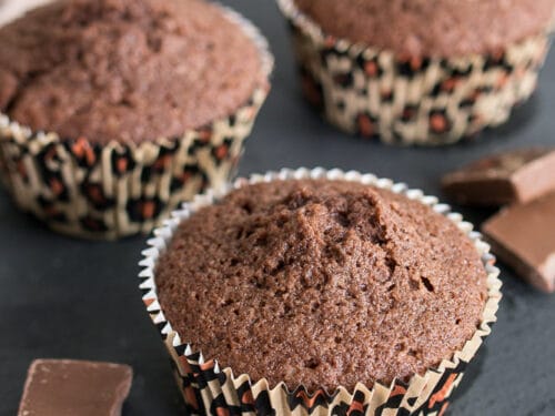 Drei Schokomuffins in Papierförmchen auf Schieferplatte mit einigen Stücken dunkler Schokolade daneben.