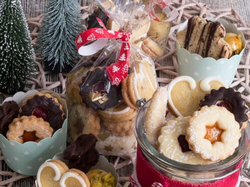 Verschiedene Weihnachtsplätzchen, darunter Vanillekipferl und mit Marmelade gefüllte Leckereien, die zusammen in Gläsern und Tassen angeordnet sind.
