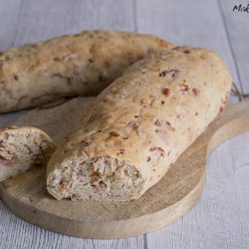 Zwei Zwiebel-Speck-Baguette mit Speckstückchen, eines davon aufgeschnitten, liegen auf einem Schneidebrett aus Holz.