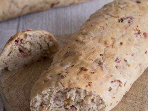Zwei rustikale Zwiebel-Speck-Baguette mit Speckstücken, eines davon angeschnitten, auf einem Holzbrett.