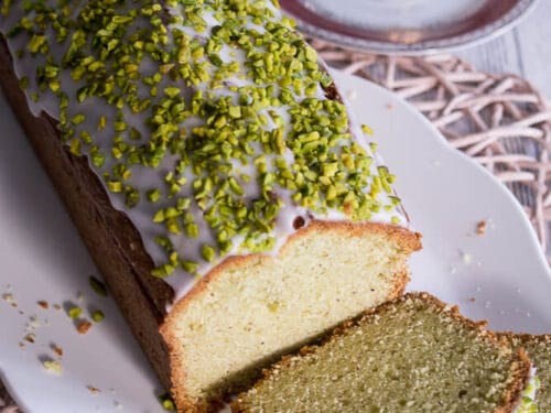 Avocado-Mandel-Kuchen aus der Kastenform mit weißer Glasur und gehackten Pistazien, in zwei Scheiben geschnitten.