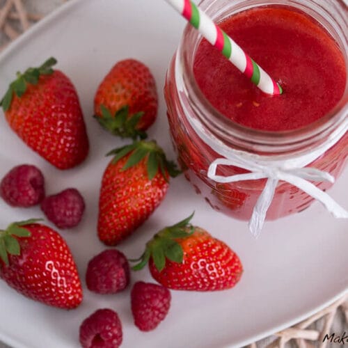 Glas mit Erdbeer- und Himbeersmoothie mit Strohhalm, umgeben von frischen Erdbeeren und Himbeeren auf einem weißen Teller.