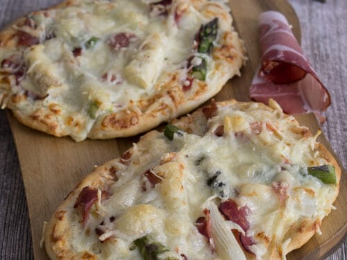 Zwei Flammkuchen mit geschmolzenem Käse, Schinken und frischem Spargel auf einem Holzbrett.