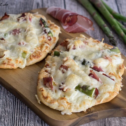 Flammkuchen mit Spargel und Schinken 3 Zwei Flammkuchen belegt mit Käse, Spargel und Prosciutto auf einem Holzbrett.
