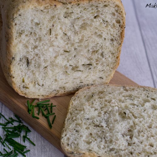 Schnittlauchbrot mit Sesam, eine abgeschnittene Scheibe liegt davor, serviert auf einem Holzbrett zusammen mit gehacktem Schnittlauch.