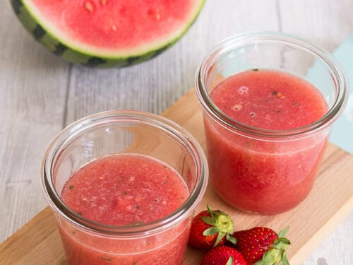 Zwei Gläser Wassermelonen-Limonade mit Erdbeeren, Minzblättern und einer halben Wassermelone auf einem Holzbrett.