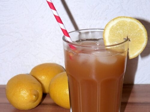 Ein Glas Eistee mit einer Zitronenscheibe und einem rot gestreiften Strohhalm, daneben drei ganze Zitronen.