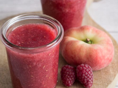 Zwei Gläser mit Himbeer-Pfirsich-Konfitüre mit einem flachen Pfirsich und zwei Himbeeren auf einem Holzbrett.