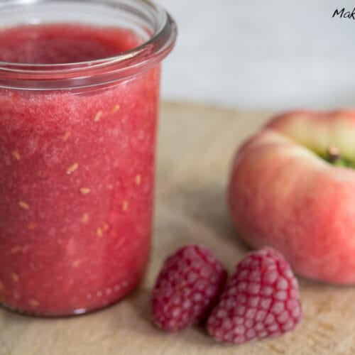 Weinbergpfirsich Marmelade mit Himbeere 3 Glas mit Himbeer-Pfirsich-Marmelade mit frischen Himbeeren und einem Weinbergpfirsich auf Holzunterlage.