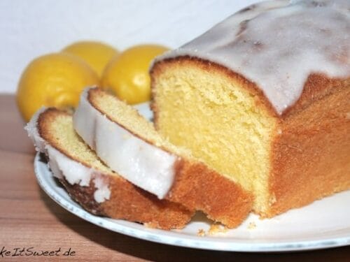 Aufgeschnittener Zitronensandkuchen mit weißem Zuckerguss auf einem Teller, begleitet von ganzen Zitronen.