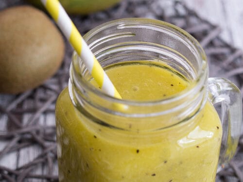 Gelber Kiwi- und Mango-Smoothie in einem Einmachglas mit gestreiftem Strohhalm, daneben frische Mango und Kiwis.