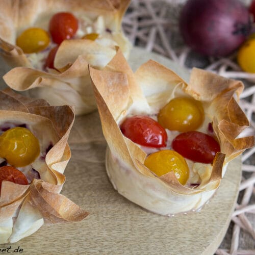 Drei mit Schafskäse und roten und gelben Kirschtomaten gefüllte Filoteigschalen auf einem Holzbrett.