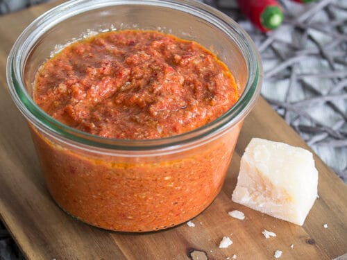 Paprika-Pesto mit Chili in einem Glas, Käsestückchen und zwei rote Chilischoten auf einem Holzbrett.