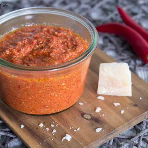 Paprika-Pesto mit Chili 3 Paprika-Pesto mit Chili im Glas, daneben ein Stück Parmesan und roten Chilischoten auf einem Holzbrett.
