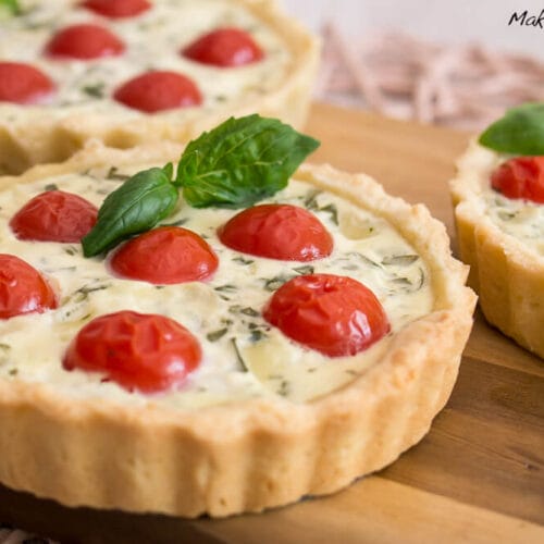 Mini herzhafte Tartelette belegt mit Kirschtomaten und Basilikum auf einem Holzbrett.