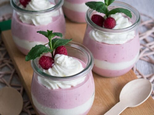 Vier Gläser mit geschichtetem rosa Himbeerquark und Sahne, Himbeeren und Minzblättern.