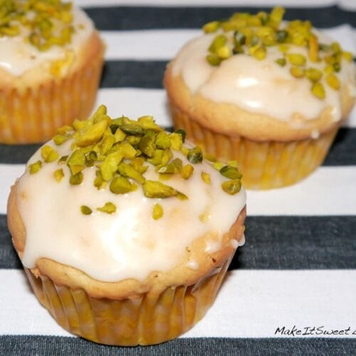 Mango Muffins mit Pistazien, Maracuja und Zitronenguss 3 Drei Mango Maracuja Muffind mit weißem Zuckerguss und gehackten Pistazien liegen auf einer gestreiften Fläche.