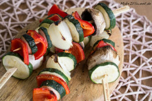 Drei gegrillte Gemüsespieße mit Zucchini, Zwiebel, roter Paprika und Champignons auf einem Holzbrett.