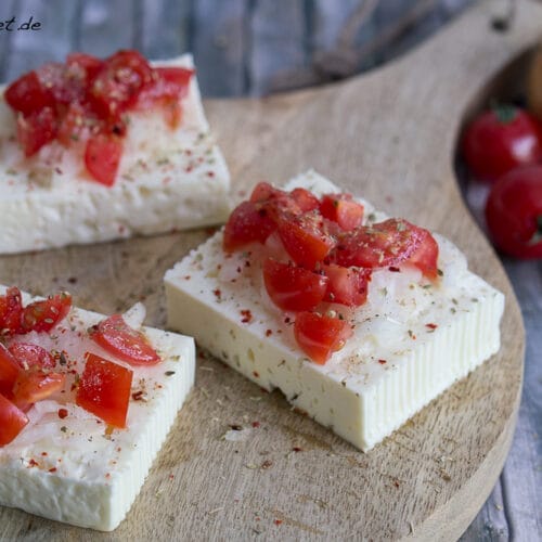 Feta mit Tomate und Zwiebeln zum Grillen 3 Feta zum Grillen, belegt mit Tomaten, Zwiebeln und Gewürzen, serviert auf einem Holzbrett.