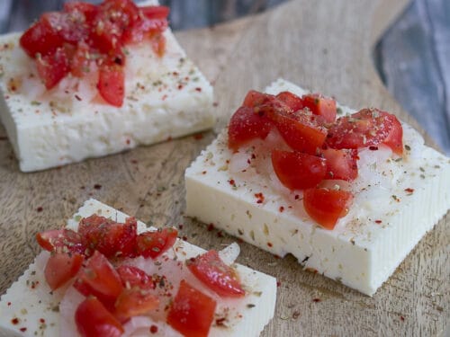 Drei Schafskäse-Scheiben, belegt mit gehackten Tomaten und Gewürzen, werden auf einem Holzbrett angerichtet.