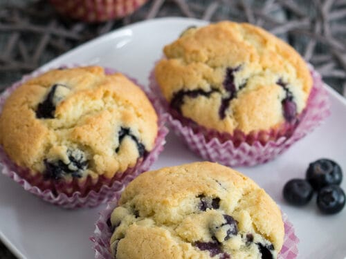 Vier schnelle Blaubeermuffins auf einem weißen Teller, ein weiterer Muffin im Hintergrund.