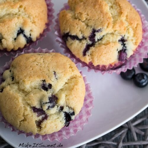 Einfache Blaubeermuffins - saftig 3 Drei einfache Blaubeermuffins in Papierförmchen auf weißem Teller mit zwei frischen Blaubeeren.