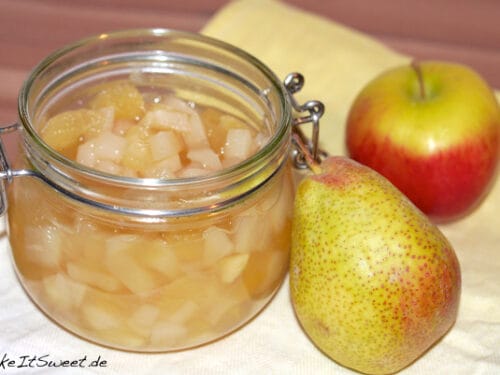 Ein Glas Vanillekompott mit Birne neben einer Birne und einem Apfel auf einem hellgelben Tuch.
