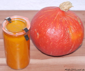 Glasgefäß mit Kürbispüree und ganzem Hokaido-Kürbis auf Holzunterlage.
