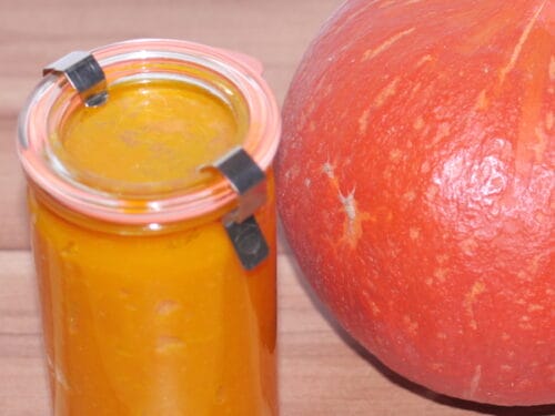 Ein Glas mit orangefarbenem Kürbispüree und ein ganzer Kürbis auf einer Holzplatte.