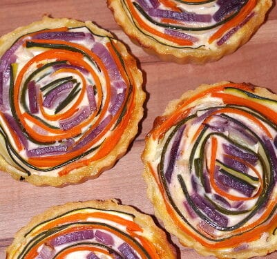 Vier Gemüsequiche-Törtchen mit Spiralen aus roter Zwiebel, Karotte und Zucchini obenauf.