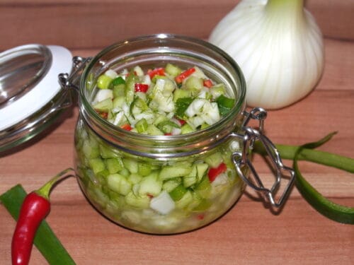 Ein Glas Gurken-Salsa mit gehackter Gurke, Zwiebel und Chili, umgeben von frischem Gemüse und einer Fenchelknolle.
