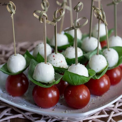 Tomate-Mozzarella-Spieße mit Kirschtomaten, Basilikumblättern und Mozzarellakugeln, hübsch auf einem weißen Teller angerichtet.