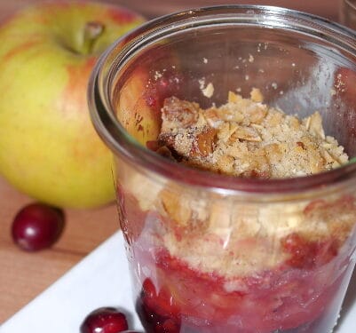 Glas mit Apfel-Cranberry-Crumble mit Streuseln auf einem weißen Teller, daneben ein ganzer Apfel.