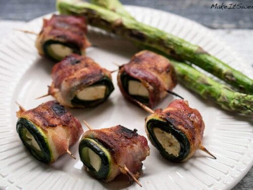 Mit Speck umwickelte Zucchini-Feta-Rollen mit geschmolzenem Käse und zwei gegrillten Spargelstangen auf einem weißen Teller.