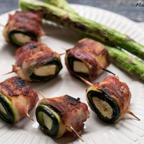 Gegrillte Zucchini Feta Röllchen mit Bacon vom Grill 3 Mit Speck umwickelte Zucchini-Feta-Rollen mit geschmolzenem Käse und zwei gegrillten Spargelstangen auf einem weißen Teller.