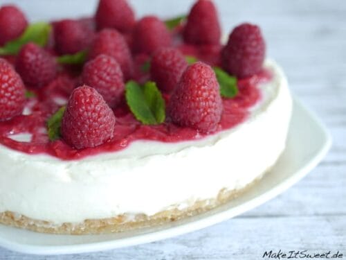Kühlschranktorte mit Himbeeren, einer weißen Creme, Keksboden, Himbeersauce und Minzblättern auf einem weißen Teller.