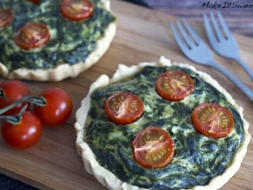 Mini-Spinat-Tartelette, belegt mit halbierten Kirschtomaten, angerichtet auf einem Holzbrett.