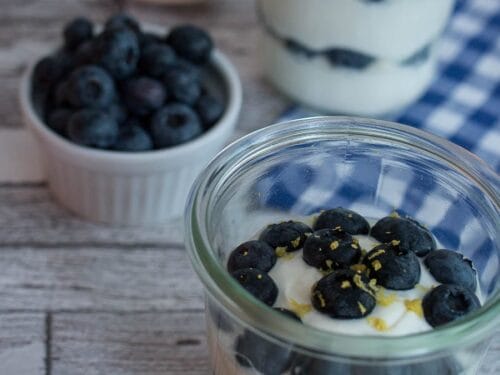 Schichtdessert im Glas mit Quark, Blaubeeren und Zitronenschale neben einer Schale mit Blaubeeren auf einem Holztisch.