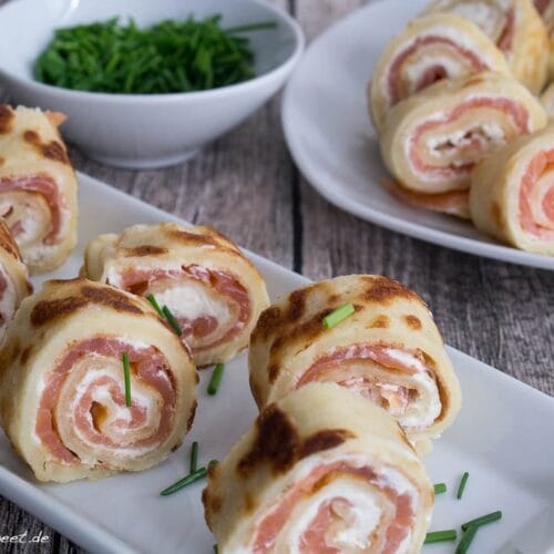 Fingerfood mit Räucherlachs Pfannkuchen 3 Pfannkuchen-Röllchen mit Frischkäse und Räucherlachs, garniert mit gehacktem Schnittlauch, serviert auf weißen Tellern.