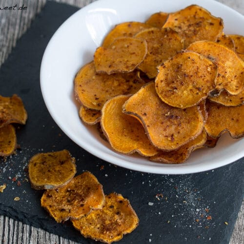 Süßkartoffel Chips selber machen 3 Gewürzte Süßkartoffelchips in einer weißen Schale, mit zusätzlichen Chips auf einer schwarzen Schieferplatte verstreut.