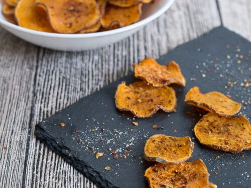 Selberstgemachte Süßkartoffelchips auf schwarzem Schiefer, mit weiteren Chips in einer weißen Schale dahinter.