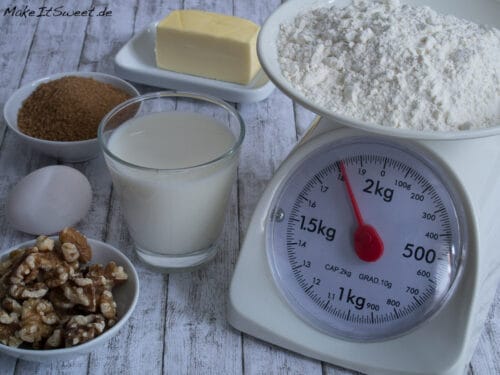 Ein Glas Milch, ein Ei, Walnüsse, brauner Zucker, Butter und Mehl auf einer Holzunterlage.