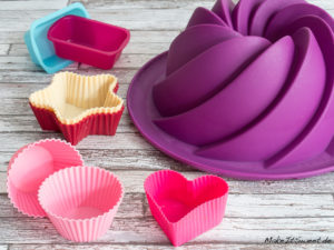 Verschiedene farbenfrohe Silikonbackformen, darunter eine große lilafarbene Gugelhupfform, die auf einer Holzfläche angeordnet sind.